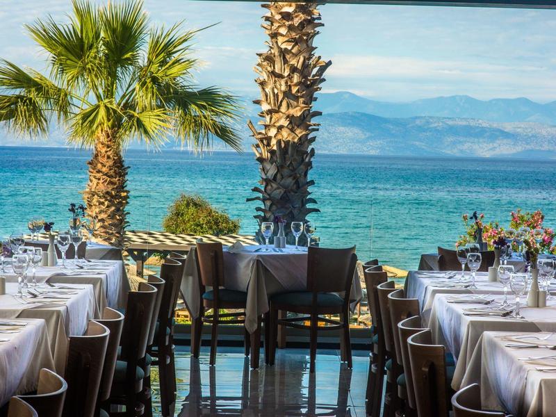 Restaurant mit gedeckten Tischen, Palmen und Blick auf das blaue Meer und Berge im Hintergrund.