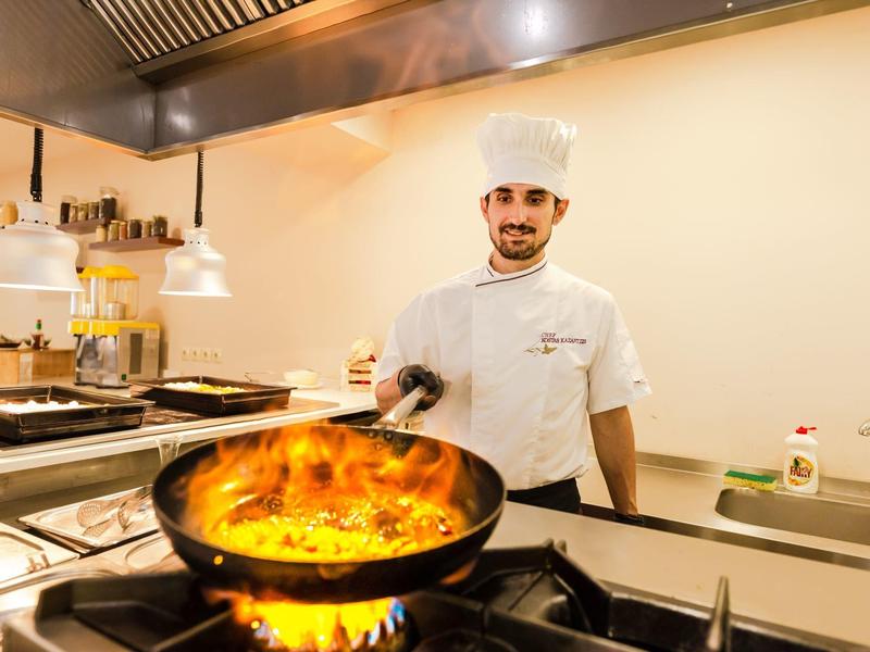 Koch in weißer Uniform steht an Herd, Pfanne mit Flammen, moderne helle Küche.