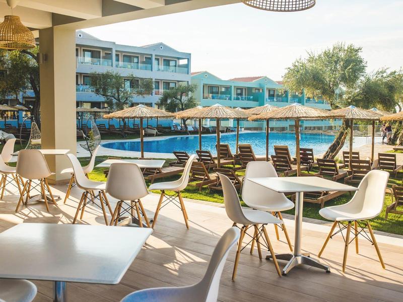 Freiluftrestaurant mit weißen Tischen, Stühlen und Blick auf einen Pool mit Liegen und blauen Sonnenschirmen.