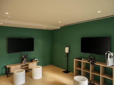 Moderner Spielraum mit zwei Fernsehgeräten, Regalen und weißen Hockern vor grünen Wänden.