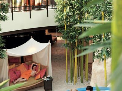 Innenraum mit Pool, drei Personen im Wasser, zwei Menschen auf Balkon, Bambuspflanzen, tropische Atmosphäre.