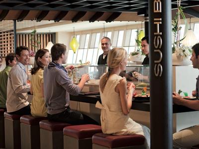 Menschen sitzen an einer Sushi-Bar, unterhalten sich und essen, helle und warme Innenraumbeleuchtung.