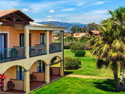 Yellow hotel building with balcony in front of green lawn and palm trees under blue sky.