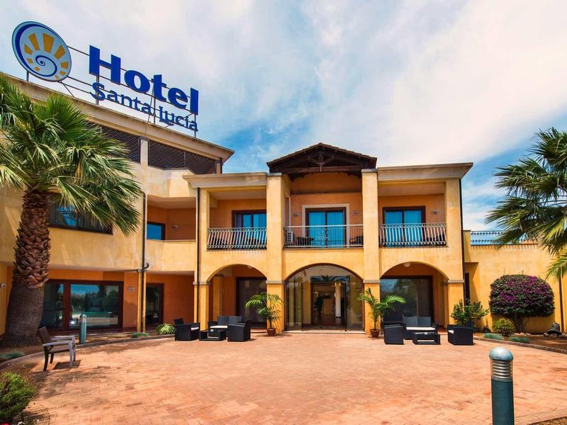 Hotel entrance with sand-colored facade, palm trees, and paved walkway.
