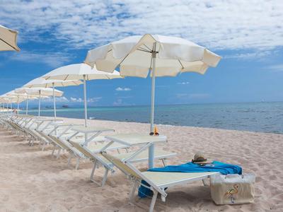 Leere Liegestühle mit weißen Sonnenschirmen am Sandstrand neben ruhigem Meer.