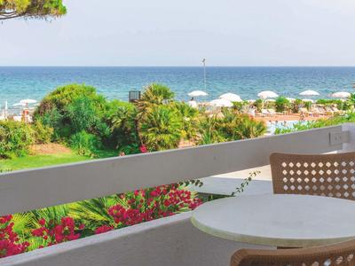 Blick von Balkon mit Tisch und Stühlen auf Garten, Strand und Meer bei klarem Himmel.