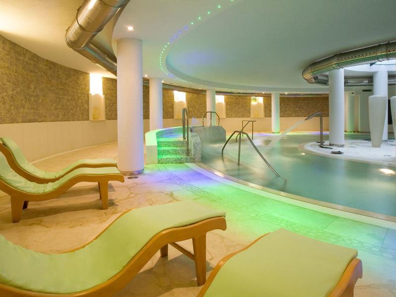 Moderner Wellnessbereich mit schimmerndem Pool, weißen Säulen und grünen Liegestühlen aus Holz.