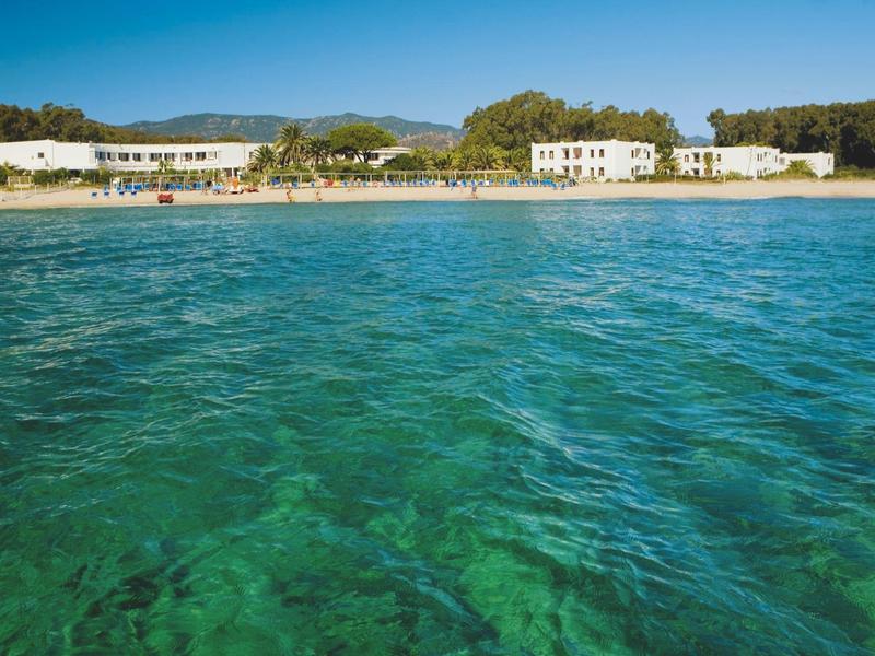Klares türkisfarbenes Meer vor einem weißen Sandstrand mit Gebäuden und grün bewaldeten Hügeln.