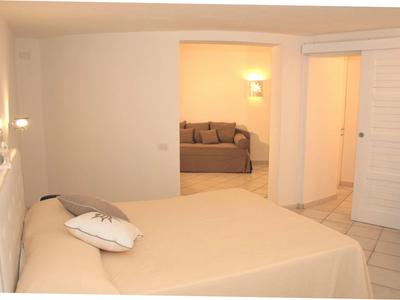 Schlichtes Schlafzimmer mit beige Bettdecke, Dekokissen, weißen Wänden und Blick ins Wohnzimmer.