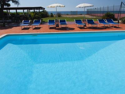 Blauer Pool mit blauen Liegen, weißen Sonnenschirmen und Bäumen im Hintergrund.