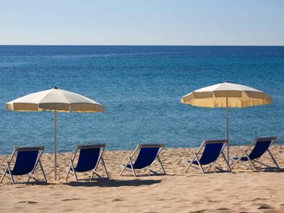 Fünf blaue Strandstühle unter zwei Sonnenenschirmen am ruhigen Sandstrand mit blauem Meer.