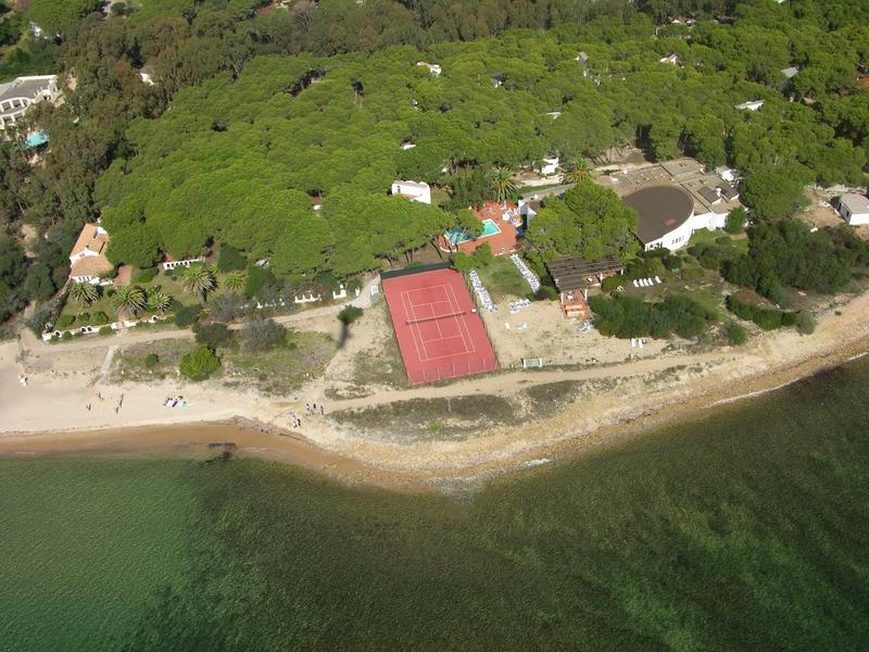 Luftaufnahme eines waldbedeckten Küstenabschnitts mit rotem Tennisplatz und grünem Meer.