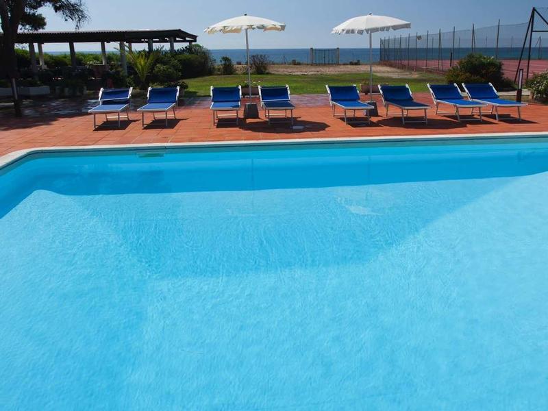 Blauer Pool mit blauen Liegen, weißen Sonnenschirmen und Bäumen im Hintergrund.