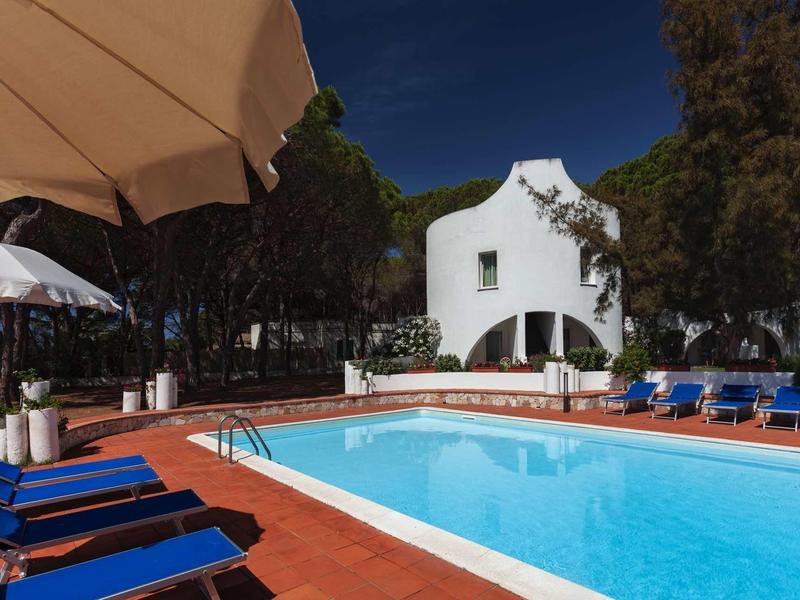 Schwimmbecken in einer Ferienanlage mit weißen Gebäuden, blauen Liegestühlen und Sonnenschirmen.