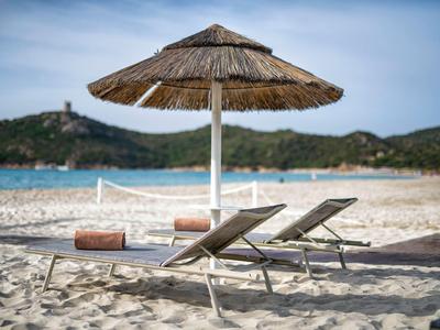 Zwei Liegestühle unter einem Strohmachschirm am Sandstrand mit Hügeln und Meer im Hintergrund.