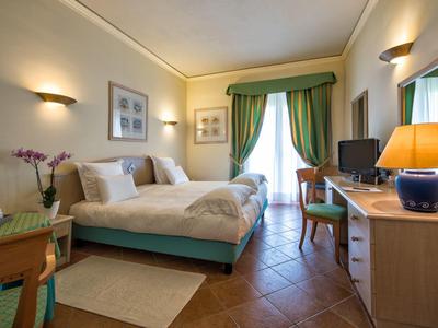 Helles Hotelzimmer mit Doppelbett, grünem Vorhang, Holzschreibtisch und dekorativer Beleuchtung.