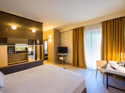 Helles Hotelzimmer mit großem Bett, TV, Schreibtisch und gelben Vorhängen am Fenster.