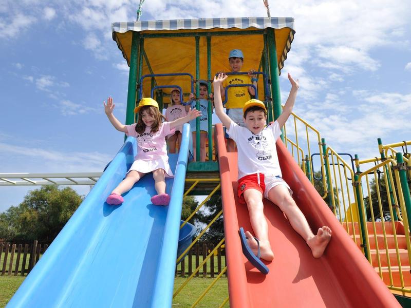 Kinder rutschen fröhlich auf blauer und roter Doppelsrutsche in sonnigem Außenbereich.