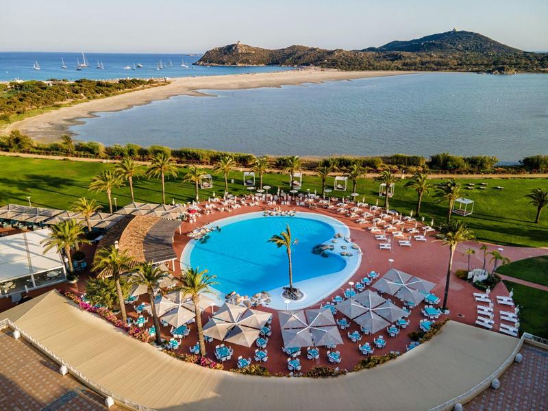 Rundförmiger Pool mit Liegestühlen und Palmen, dahinter Sandstrand und hügelige Landschaft.