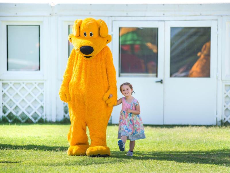 Ein kleines Mädchen hält die Hand einer großen Person im gelben Hundekostüm auf einem Rasen vor einem Haus.
