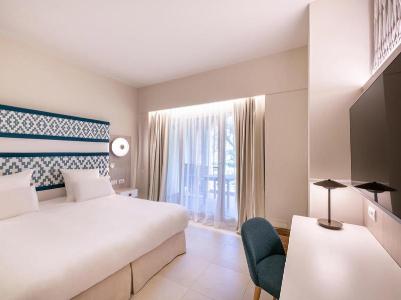 Modernes Hotelzimmer mit großem Bett, Schreibtisch, Sessel und hellem Vorhang an Fensterfront.