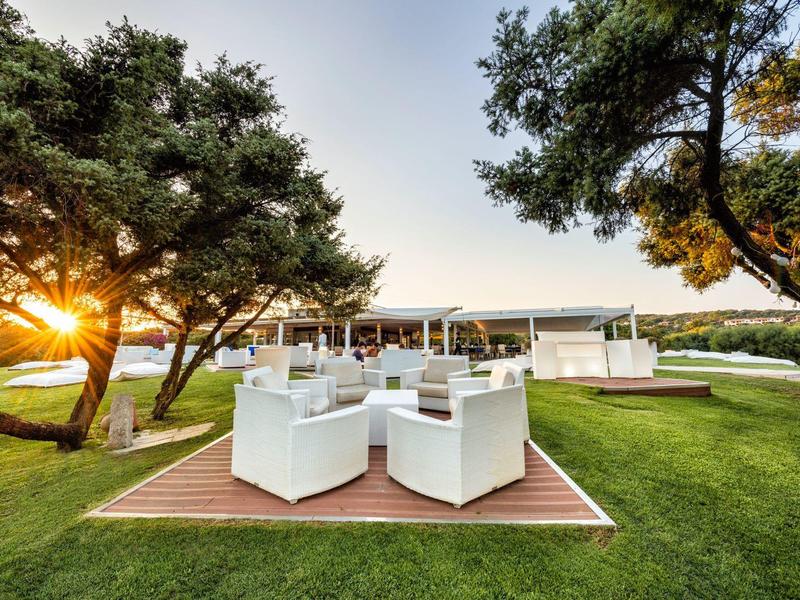Garten mit weißen Möbeln auf Holzdeck, grüner Wiese und Bäumen bei Sonnenuntergang.