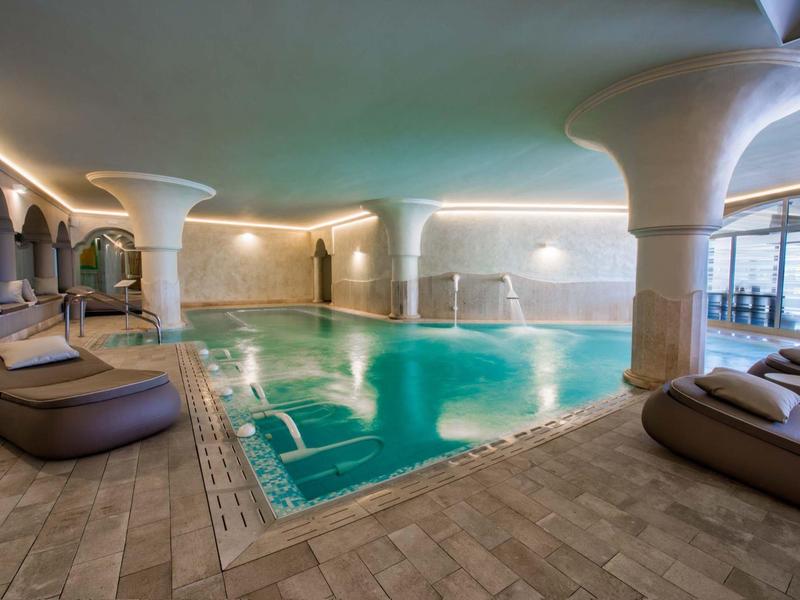 Innenpool mit Säulen, warmem Licht, Liegen und Mosaikfliesen am Boden und an den Wänden.