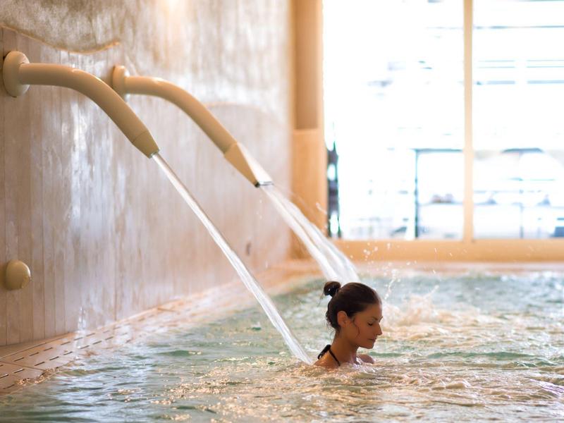 Frau entspannt im Hallenbad mit Wasserstrahlen aus zwei Rohren an der Wand.