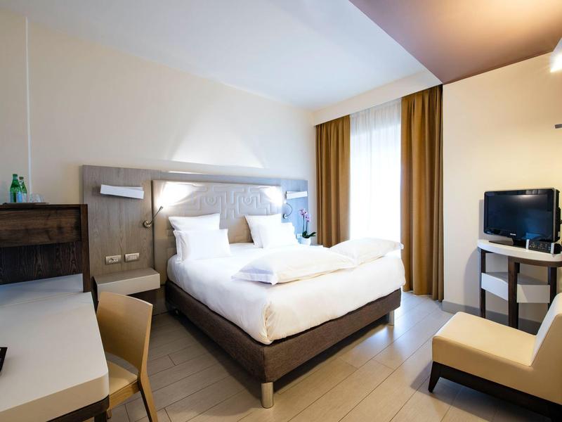 Helles Hotelzimmer mit Doppelbett, Schreibtisch, Fernseher und bequemen Stuhl, moderne Ausstattung.