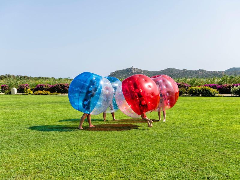 Menschen spielen Bubble Soccer auf grünem Rasen, umgeben von Büschen und Bergen im Hintergrund.
