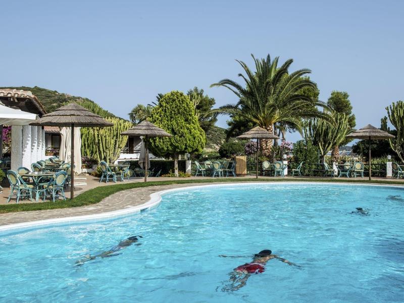 Zwembad met zwemmers, omgeven door parasols, palmbomen en kleine cottages onder een blauwe lucht.