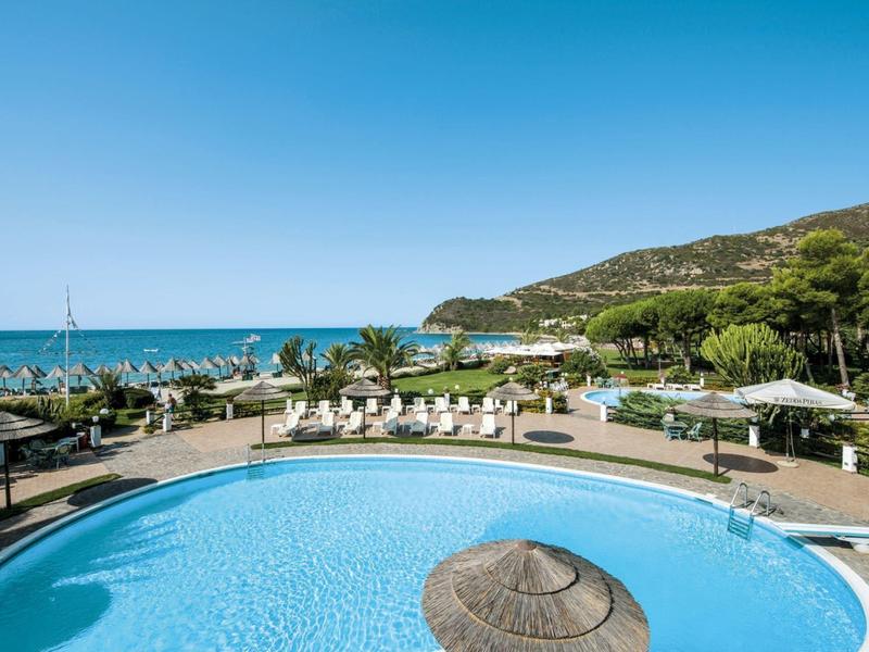 Rond hotelzwembad met parasols en uitzicht op zee en groene heuvels onder een heldere hemel.