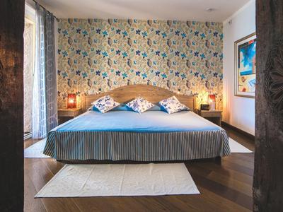 Cozy bedroom with double bed, patterned pillows and wallpaper, wooden floor and ample light.