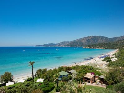 Coastline with clear blue water, sandy beach, green vegetation, and mountains in the background.