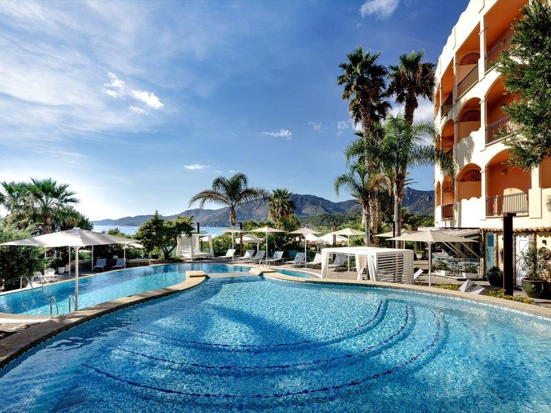 Large outdoor pool next to a hotel with palm trees and mountain and sea views.