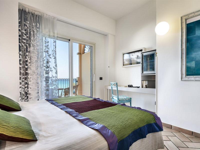 Brightly furnished bedroom with double bed, desk, and balcony with view.