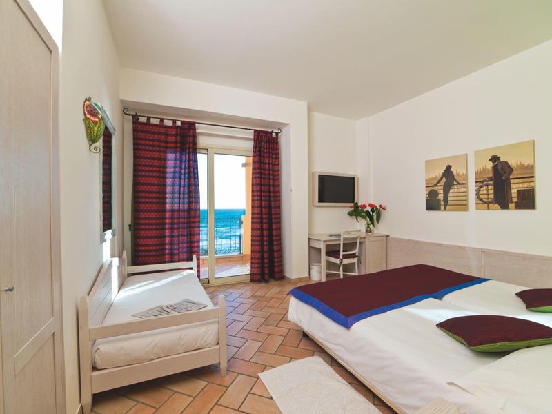 Bright hotel room with double bed, sofa, desk, and sea view through window with red curtains.