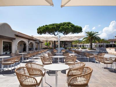 Moderne buitenruimte van een hotel met rieten meubilair, tafels en een fontein onder een blauwe lucht.