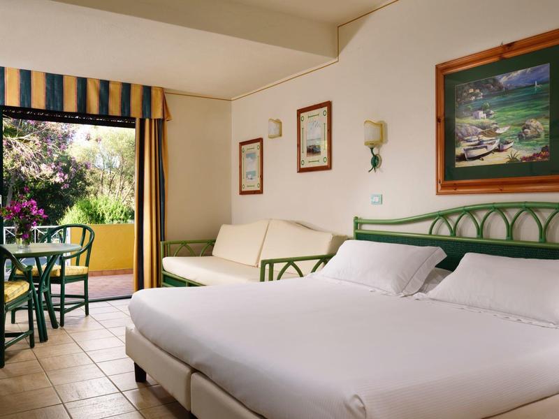 Licht hotelkamer met tweepersoonsbed, bank, eettafel en balkon met groen uitzicht.