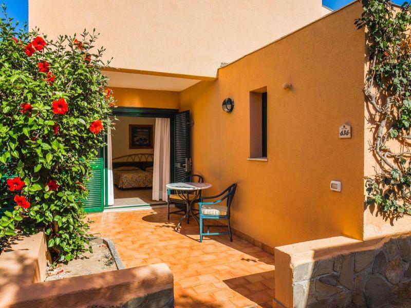 Ingang van een geel vakantieappartement met terras en bloemen bij zonnig weer.