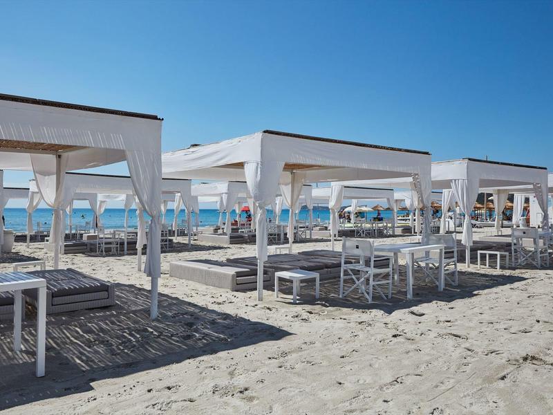 Meerdere witte strandpaviljoens met gordijnen en tafels op zand onder een blauwe lucht.