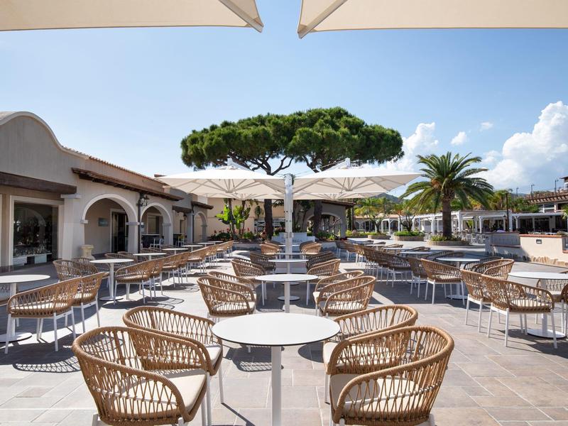 Moderne buitenruimte van een hotel met rieten meubilair, tafels en een fontein onder een blauwe lucht.