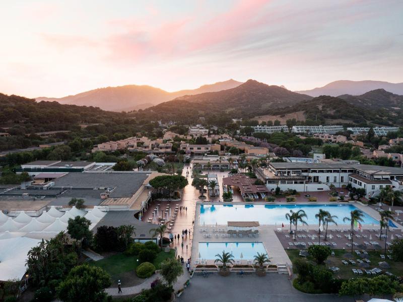 Uitzicht op een resort met zwembaden en palmbomen bij zonsondergang in een bergachtig landschap.