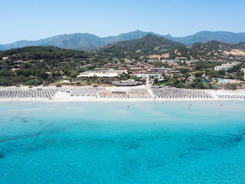 Helder turquoise zee voor een breed zandstrand met parasols en heuvels met begroeiing erachter.
