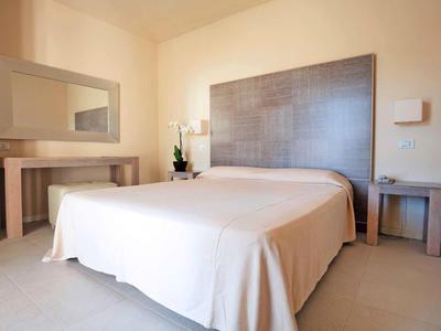 Jasny pokój hotelowy z dużym łóżkiem, stolikami nocnymi, lustrem i prostą dekoracją.