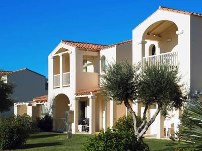 Weiße mediterrane Ferienhäuser mit roten Dächern und gepflegtem Garten unter blauem Himmel.