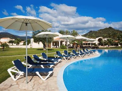 Duży basen z niebieską wodą, białymi parasolami i leżakami przed domkami wakacyjnymi.