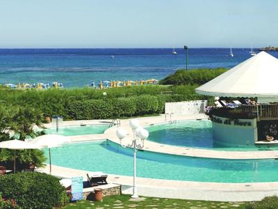 Pool mit Rundbar, Liegestühlen und grüner Bepflanzung am Meer bei klarem Himmel.