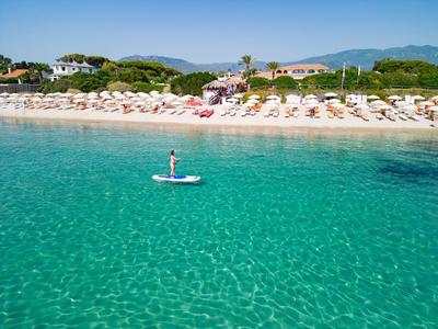 Klares türkisfarbenes Meer mit Person auf Paddleboard, weißer Sandstrand und Sonnenschirme im Hintergrund.