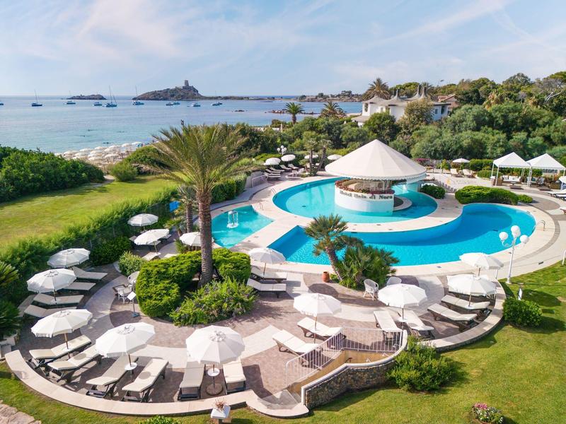 Runder Pool mit weißem Sonnenschirm und Liegestühlen in grüner Gartenanlage am Meer.